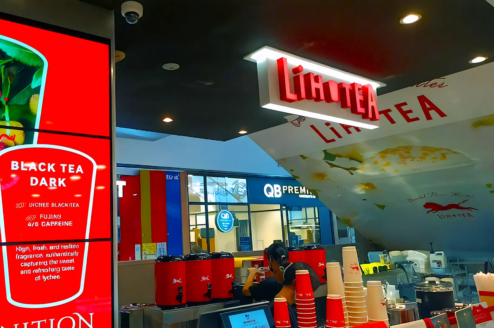 A vibrant tea shop with a red sign reading "Lihotea," features a digital menu promoting "Black Tea Dark." An employee behind the counter prepares drinks.