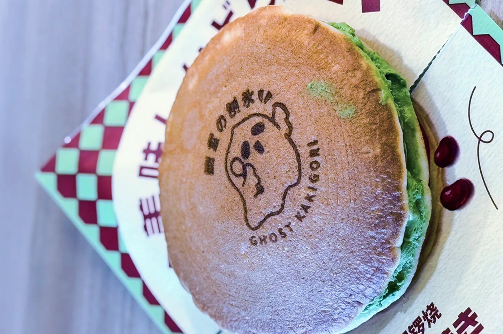 A round pancake engraved with a cartoon ghost and text "Ghost Taiyaki," filled with green filling, on checkered packaging. Whimsical and playful mood.