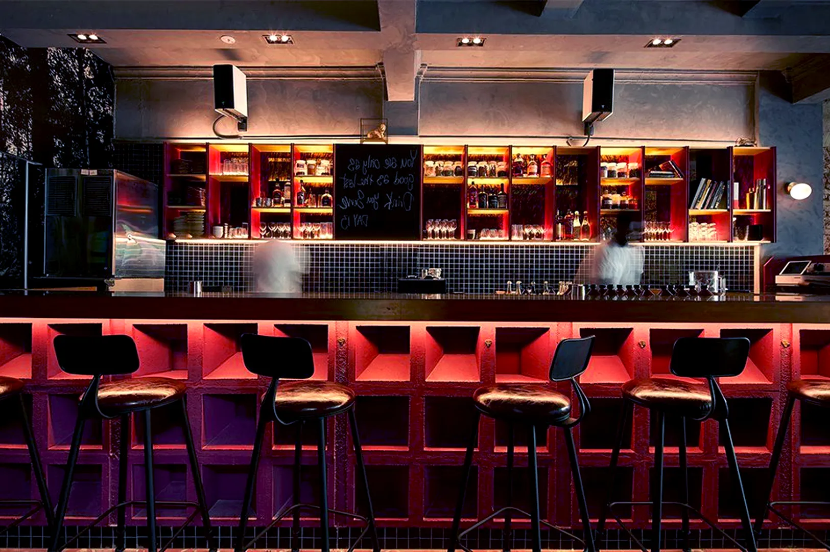 Dimly lit bar interior with a warm, cozy ambiance. Five high stools line the bar, which is backlit in red. Shelves hold glasses and bottles.