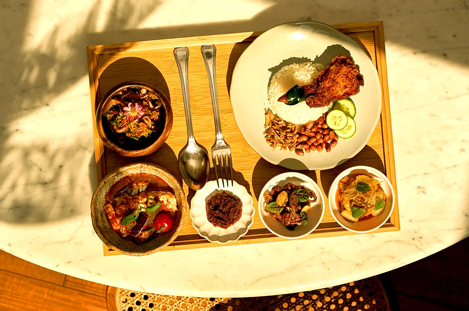 A wooden tray with a Malaysian meal on a marble table. Includes rice, fried chicken, peanuts, cucumber, sambal, two curry bowls, utensils, and sunlight.