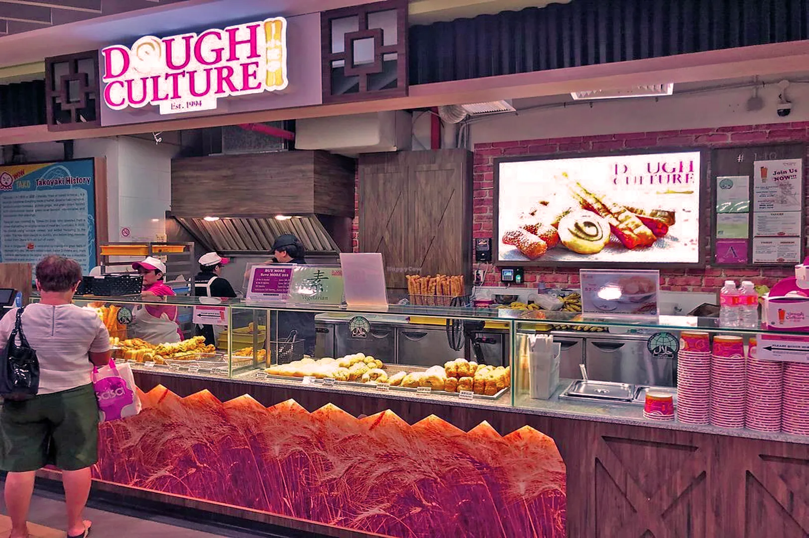 A woman stands in front of the Dough Culture food stall, lined with pastries in glass displays. The vibrant setting conveys a bustling atmosphere.