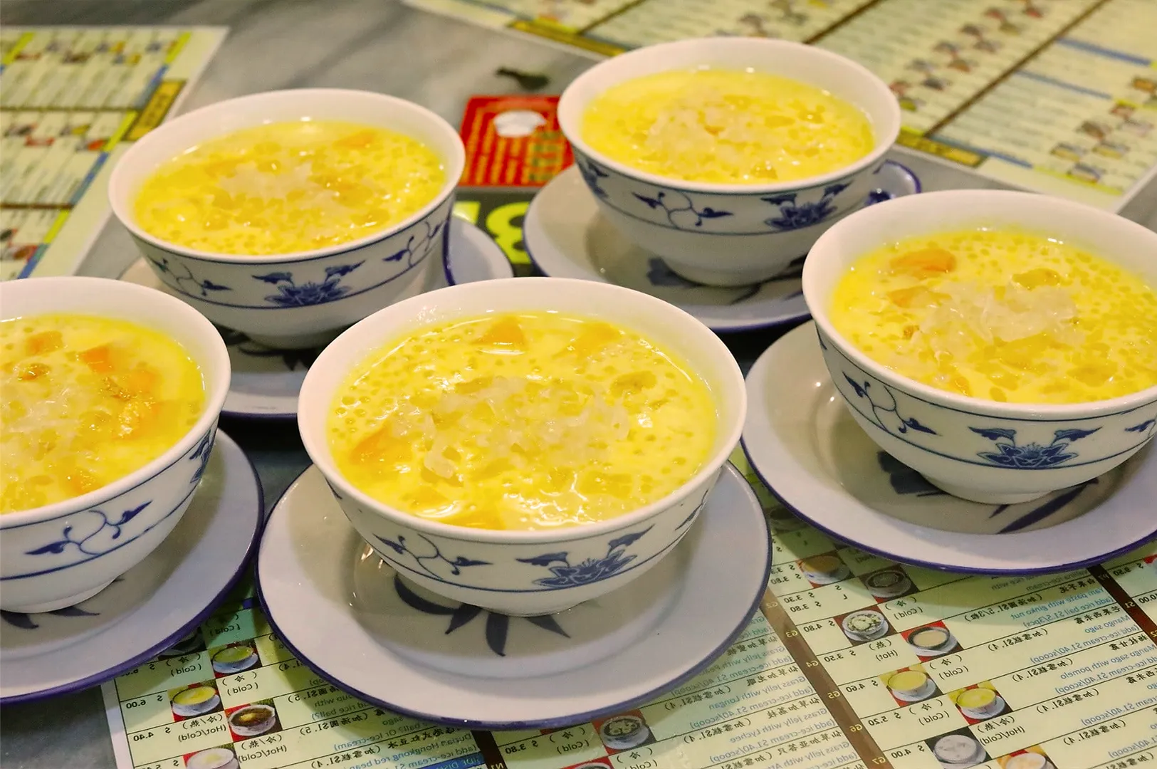 Five bowls of mango sago dessert with shaved ice, served in blue and white patterned bowls on saucers, arranged on a table with menus.
