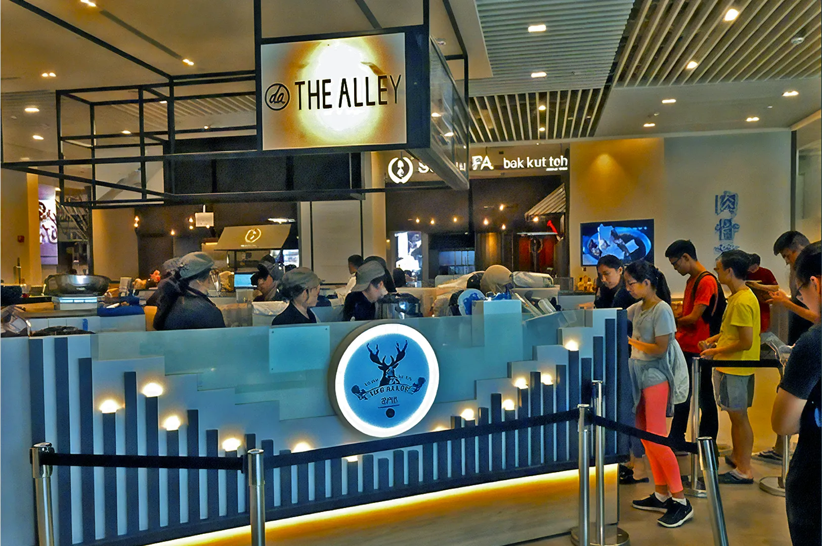 A bubble tea shop with a sign reading "The Alley" features a modern, illuminated counter with a deer logo. Customers line up, creating a lively atmosphere.