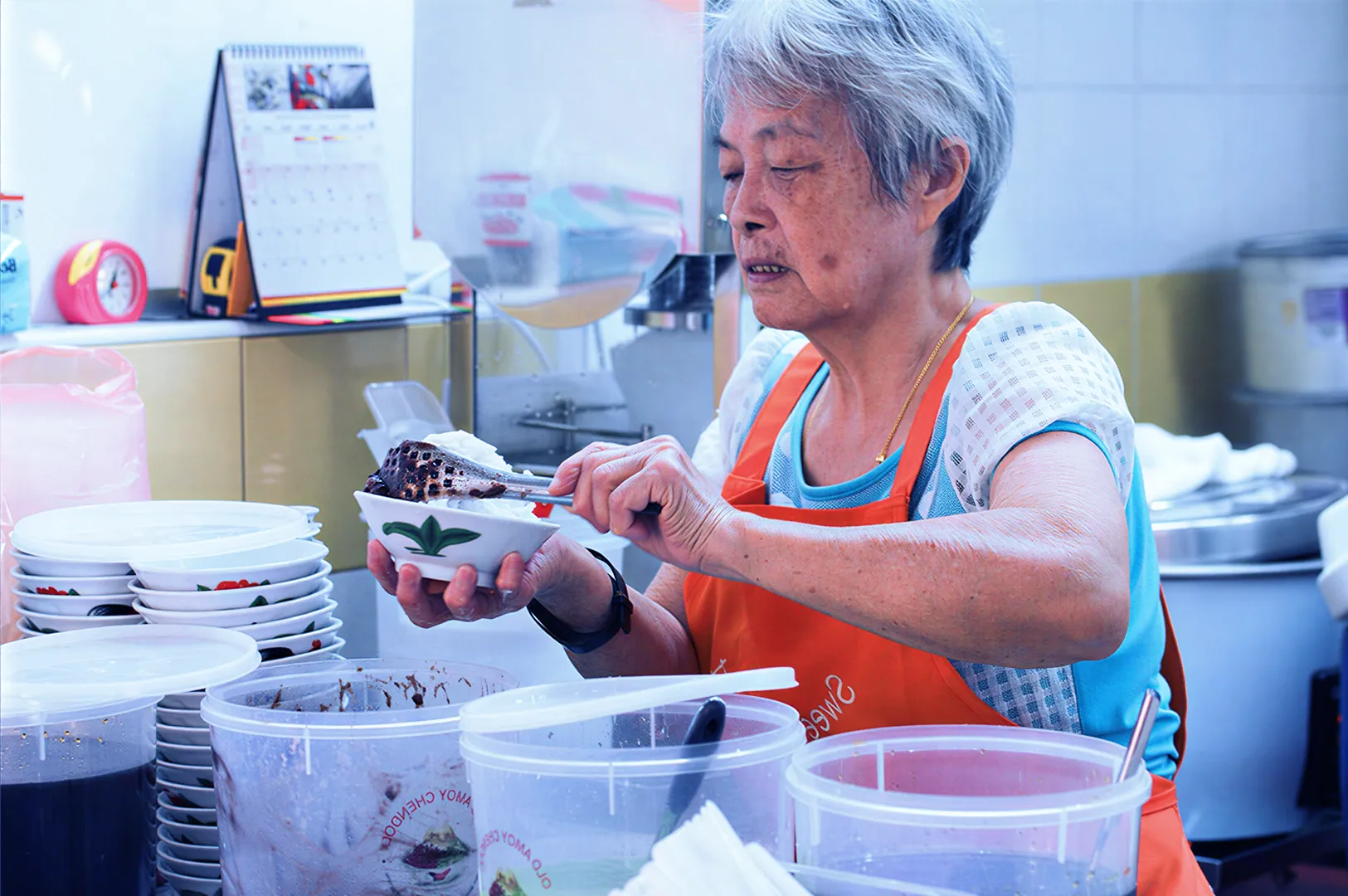 Elderly woman in an apron serves food into a bowl at a busy kitchen counter. Surrounding her are stacked bowls and containers, creating a lively, focused atmosphere.
