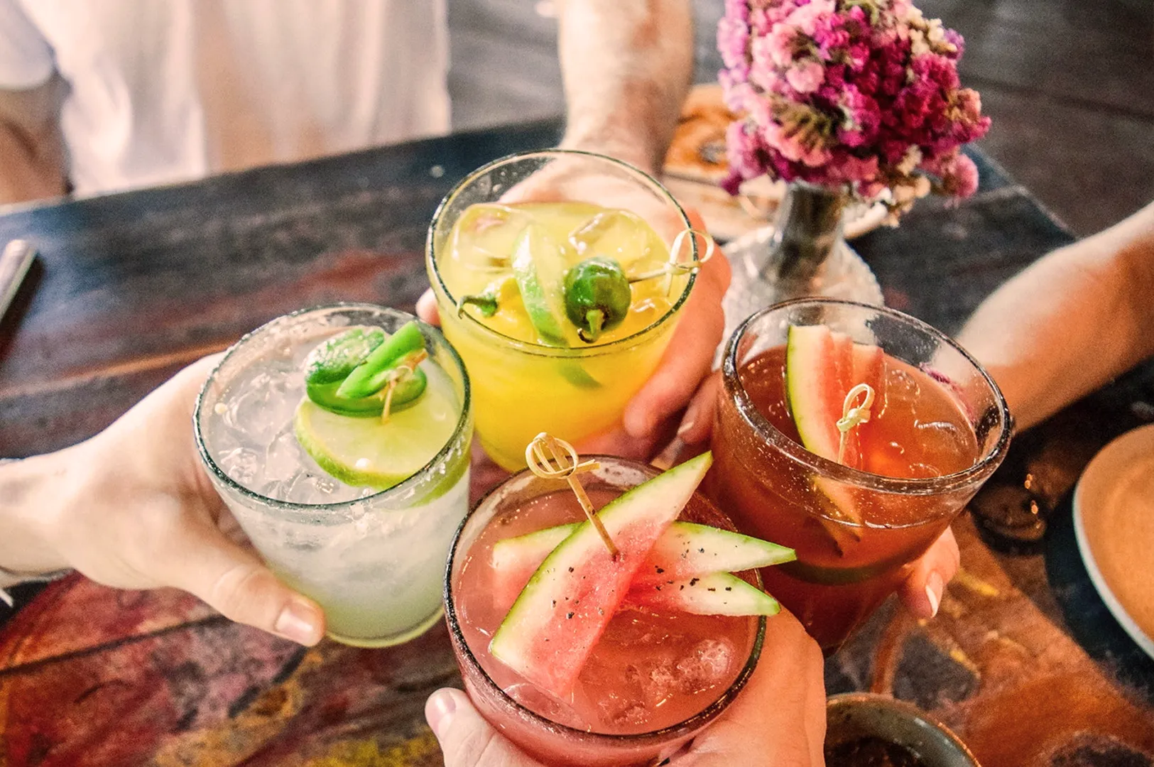 Hands holding four colorful cocktails garnished with fruit and jalapeños over a wooden table. A small vase of purple flowers sits nearby, creating a festive mood.