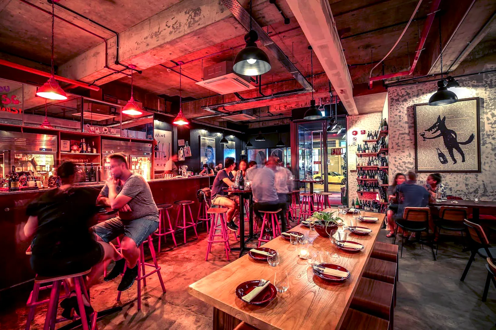 A dimly lit urban bar with exposed pipes and red lighting. Patrons converse on high stools by the bar. A wooden table with plates and glasses in the foreground adds warmth. A wolf-themed artwork hangs on a textured wall. Cozy, lively ambiance.