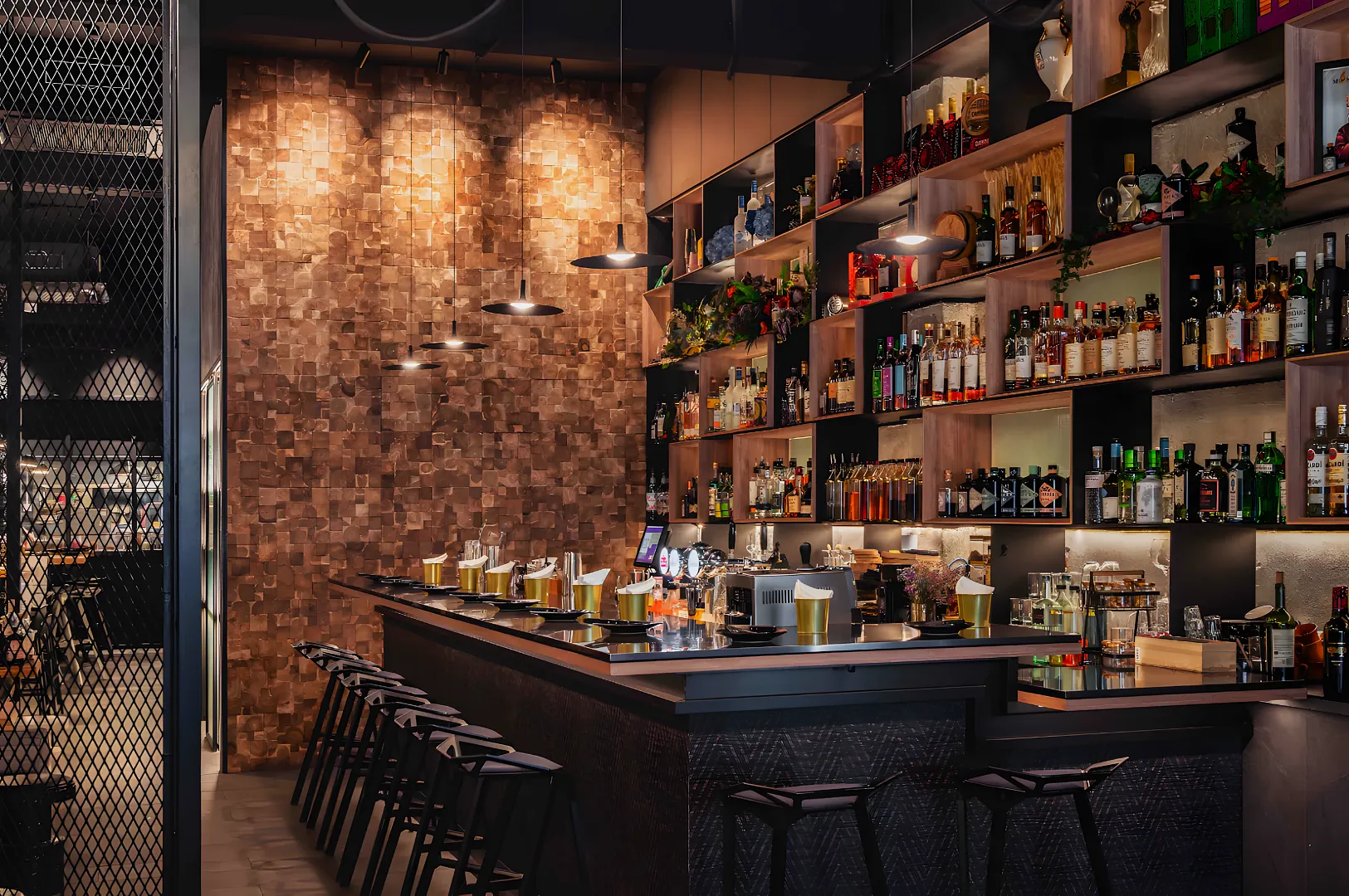 Cozy bar interior with wooden shelves stocked with assorted liquor bottles. Warm lighting casts a soft glow over the counter lined with barstools.