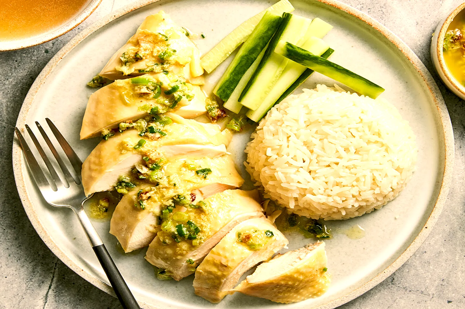 Sliced poached chicken with sauce, cucumber sticks, and a mound of rice on a plate. A fork rests on the side. The setting is calm and inviting.