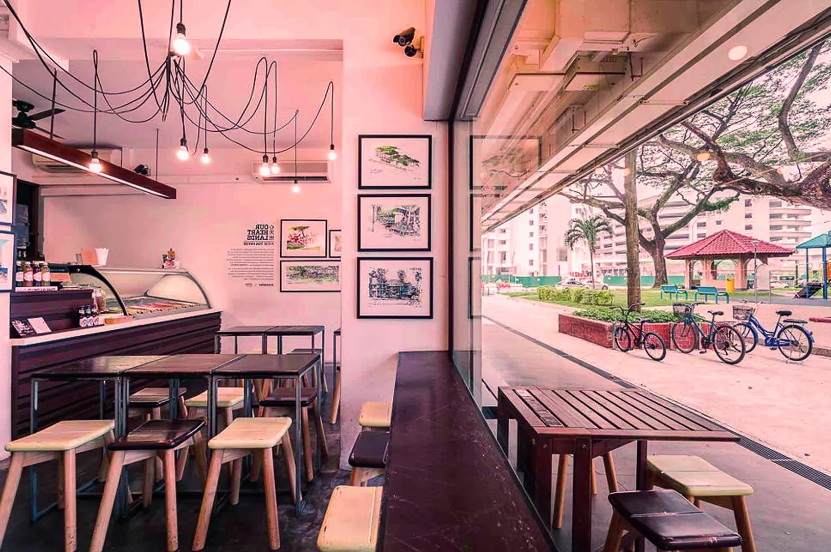 Cozy cafe interior with wooden tables and stools, adorned with framed artwork. Large windows reveal a scenic street view with parked bicycles and trees. Warm, inviting ambiance.