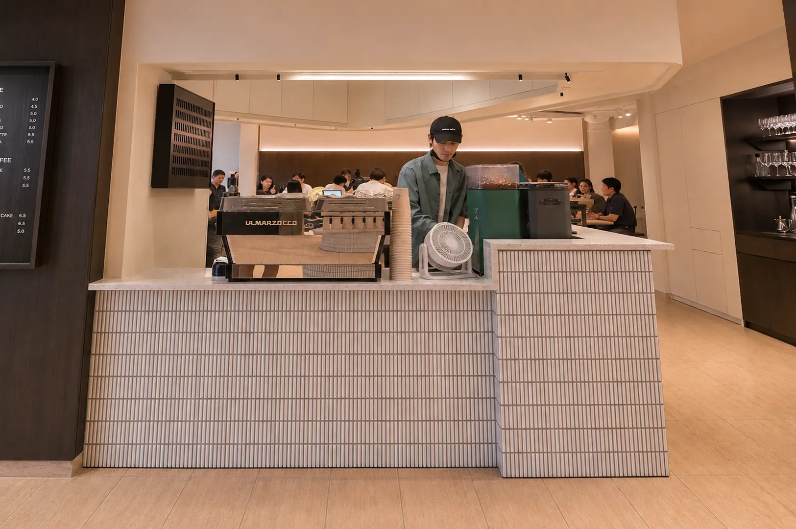 Modern coffee shop counter with barista in black cap working behind white tiled counter, La Marzocco espresso machine, stacked cups, menu board on wall, and customers seated in background