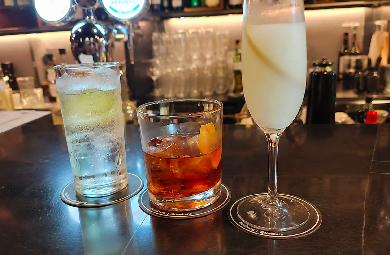 Three cocktails on a bar counter: a highball, an old fashioned, and a tall white drink.