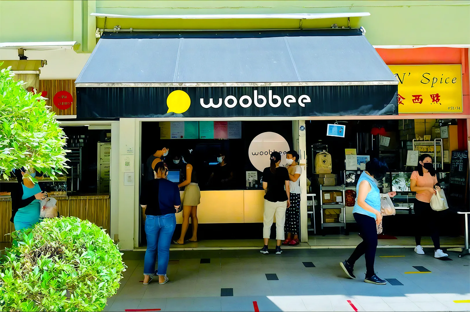 People stand in line outside a shop with a black awning labeled "woobbee." The scene is sunny, lively, and includes greenery in the foreground.