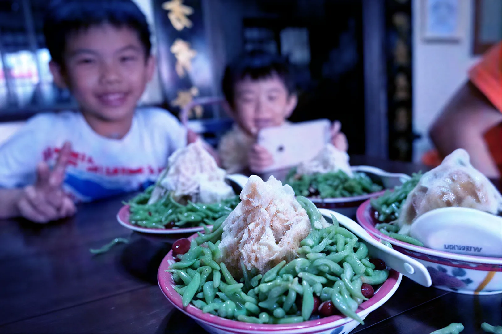 Two children joyfully sit at a table with bowls of green cendol topped with ice and syrup. One child makes a peace sign, while the other holds a phone.