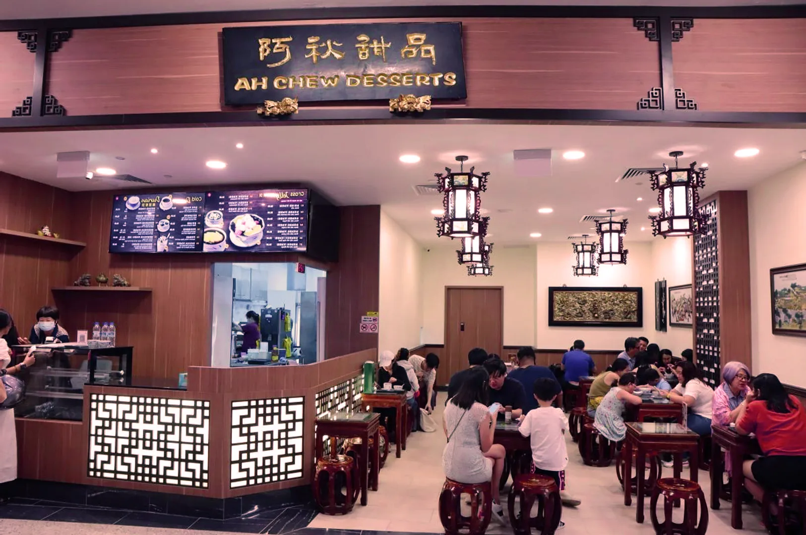 Cozy dessert shop with Asian decor, ornate lighting, and a wooden sign reading "Ah Chew Desserts." Patrons enjoy treats at round tables.