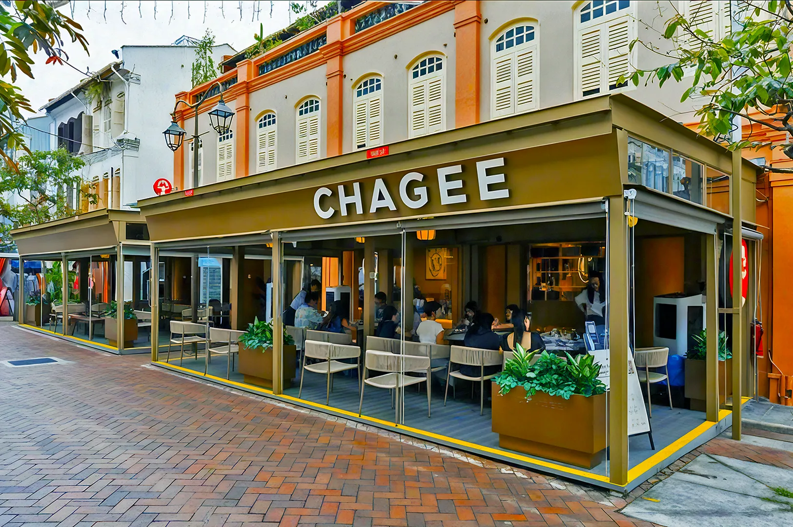 Outdoor view of "Chagee" cafe on a vibrant street corner, featuring modern seating and a bustling, inviting atmosphere with people inside.