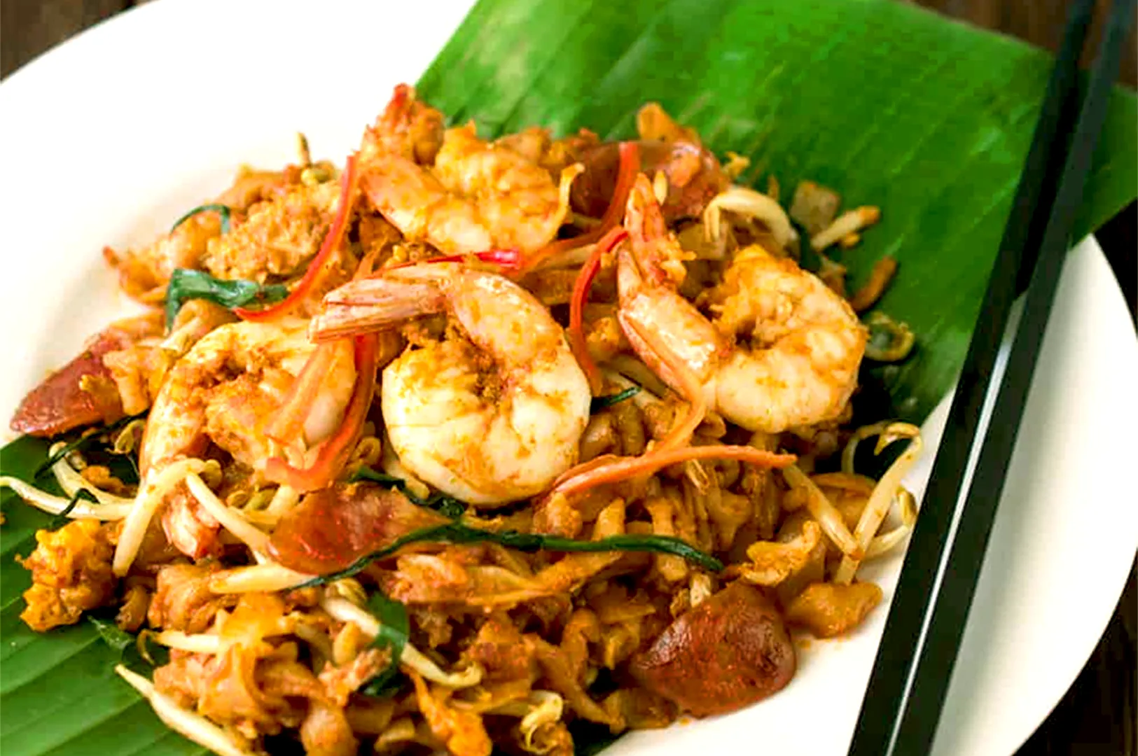 A plate of char kway teow on a banana leaf, featuring shrimp, noodles, bean sprouts, and chili, with chopsticks on the side, evoking a savory and spicy appeal.