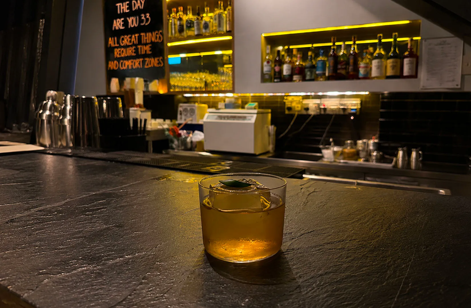 A single amber cocktail on a dark bar counter with warm backlighting.
