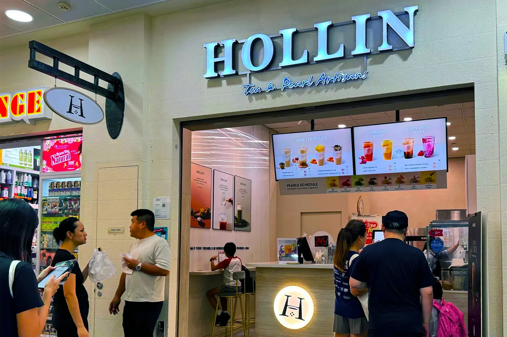 Storefront of "Hollin Tea & Pearl Artisans." Customers line up inside. Menu displays various drinks. Bright, inviting atmosphere with a modern design.