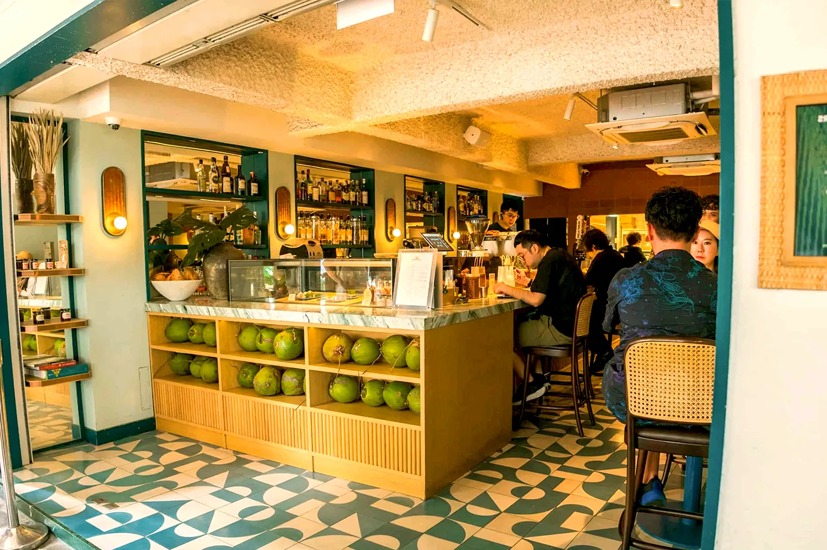 Cozy cafe interior with a tropical theme, featuring a counter lined with fresh coconuts, colorful floor tiles, and patrons enjoying drinks at the bar.
