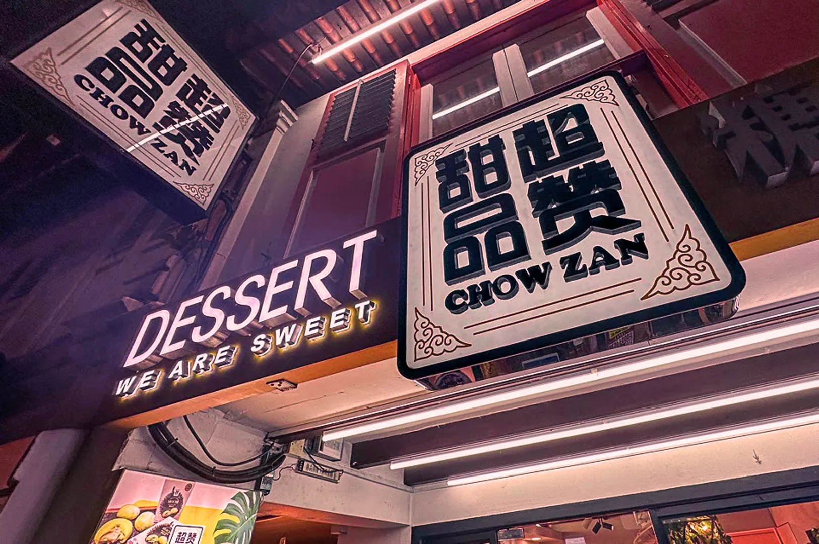 Storefront with two illuminated signs at night. One reads, "DESSERT WE ARE SWEET," the other features Chinese characters and "CHOW ZAN". Warm lighting creates a cozy atmosphere.