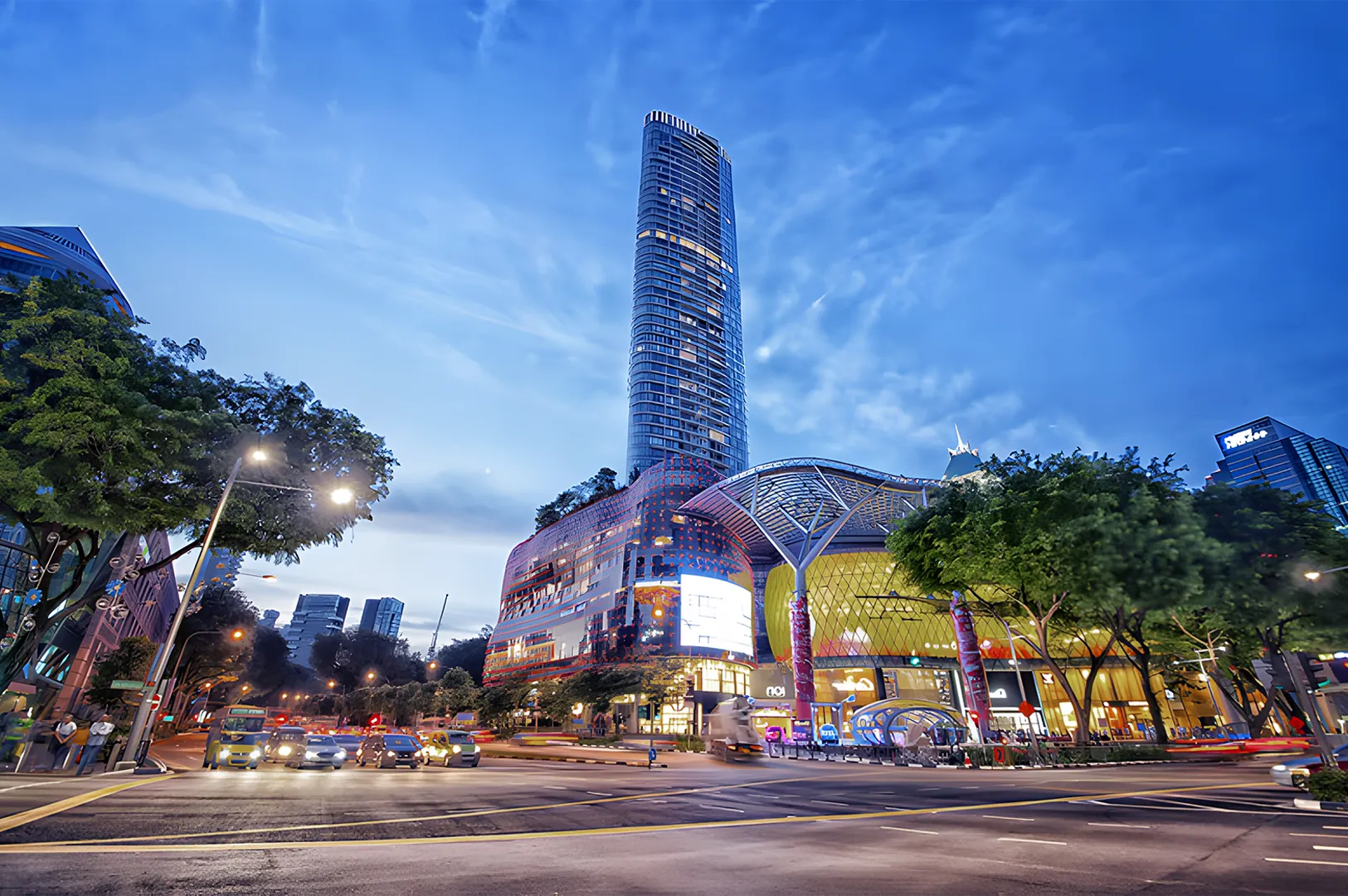 Tall skyscraper and modern shopping mall under a vibrant evening sky, surrounded by busy streets and cars, creating a lively urban atmosphere.
