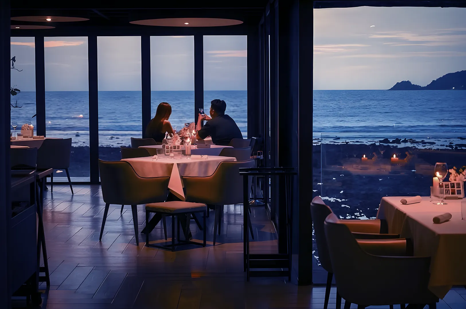 A couple dines at a candlelit restaurant with large windows overlooking a serene ocean at dusk. The atmosphere is intimate and tranquil.