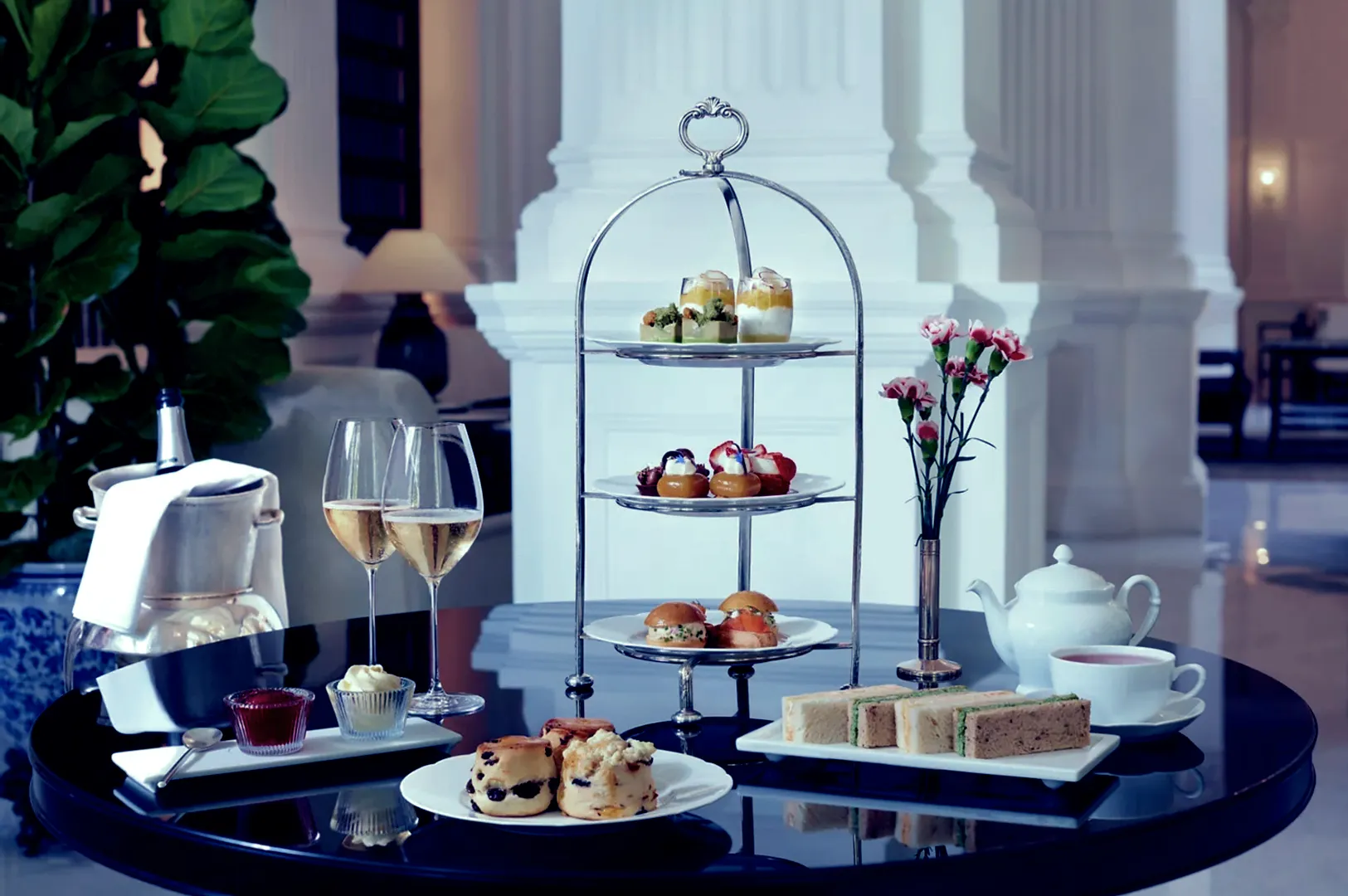 Elegant afternoon tea setting with a three-tiered stand of pastries and sandwiches, champagne glasses, a teapot, and fresh flowers on a glossy table.