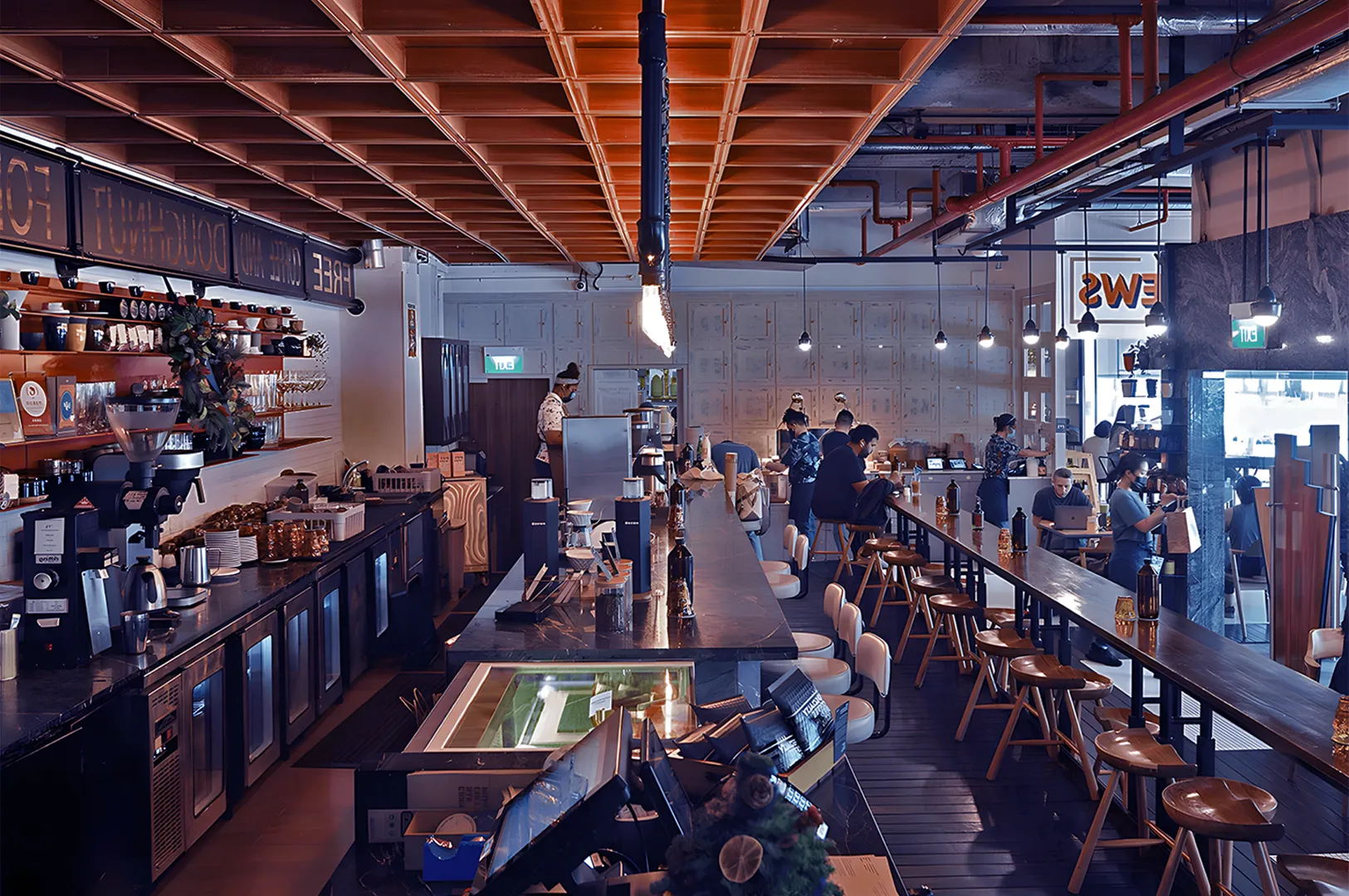 A bustling modern café with high ceilings, bar seating, and warm lighting. Patrons are seated along a long counter and tables, creating a lively atmosphere.