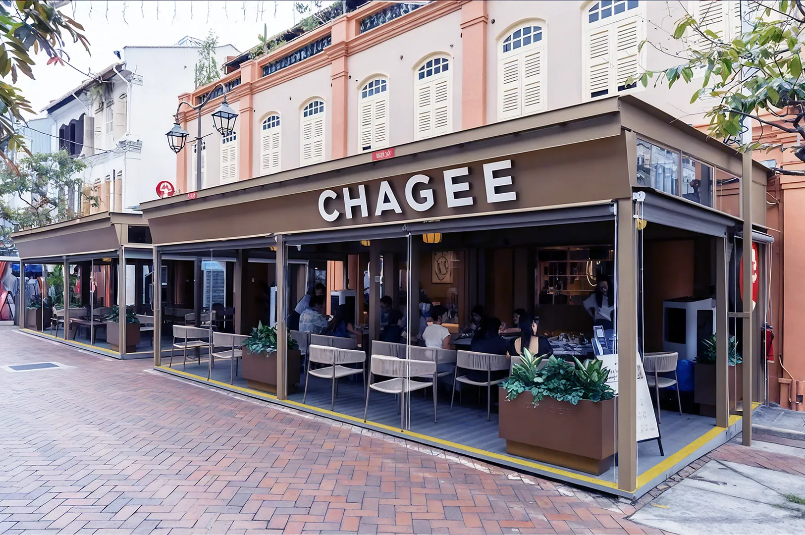 Outdoor view of "Chagee" tea house with people seated at tables under a canopy. The building has a classic style, creating a cozy atmosphere.