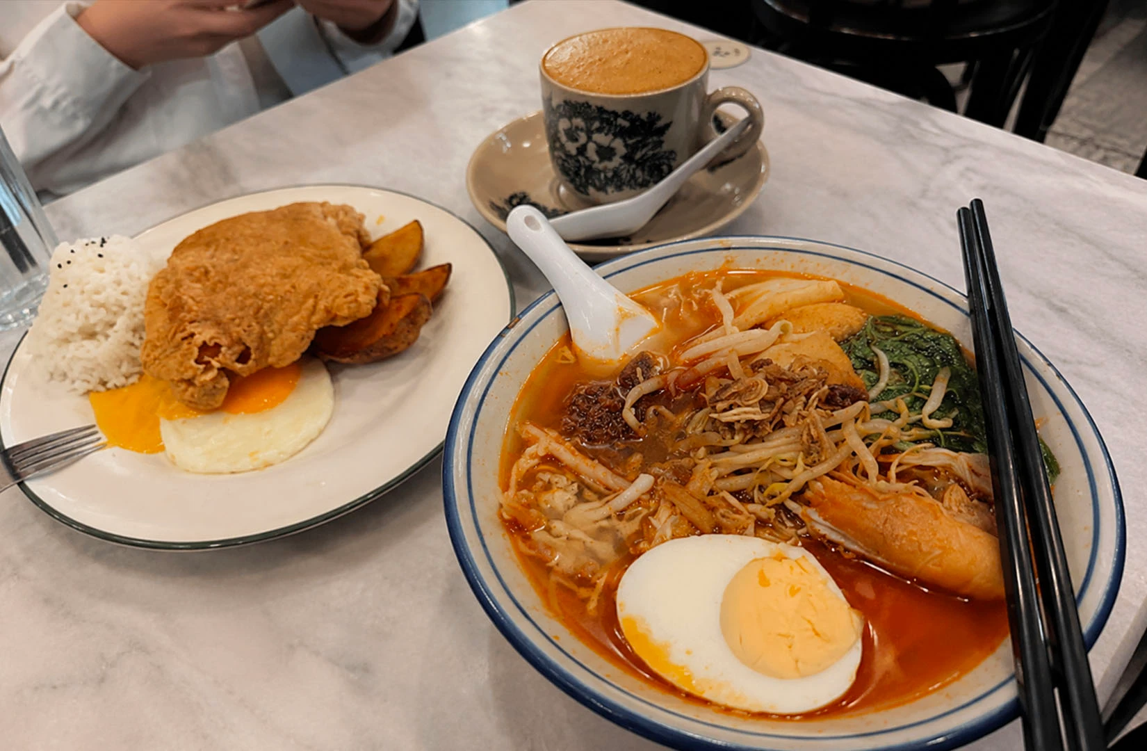 A marble table displays a bowl of spicy noodle soup, a plate of fried chicken chop with rice, and a cup of traditional kopi.i
