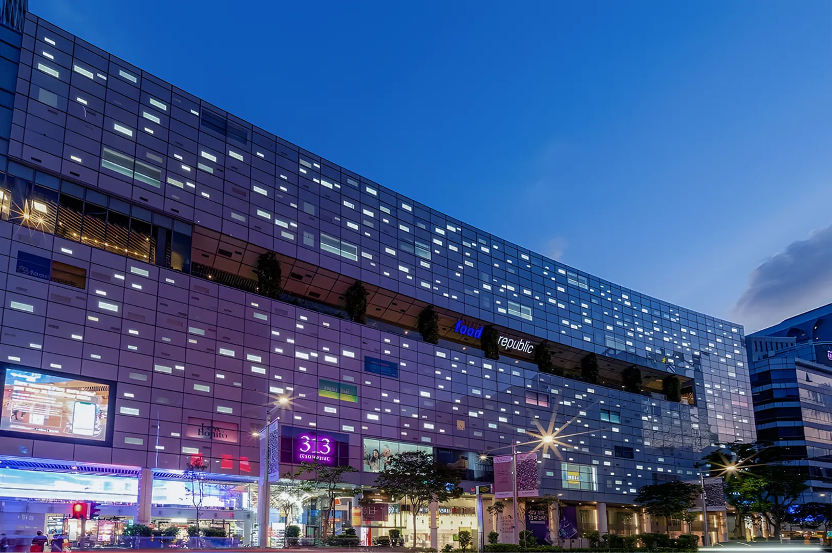 Modern shopping mall facade at dusk, illuminated by glowing windows and signs, including "Food Republic," against a clear evening sky.