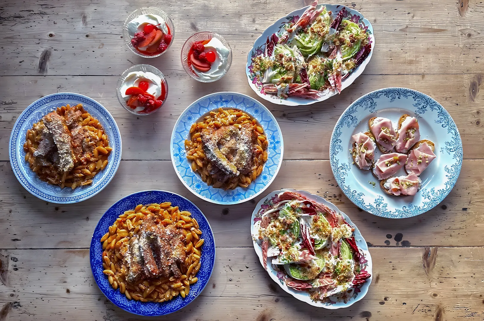 A rustic wooden table with three pasta dishes topped with crispy meat, two salads, sliced cured meat on a plate, and three bowls of yogurt with strawberries.