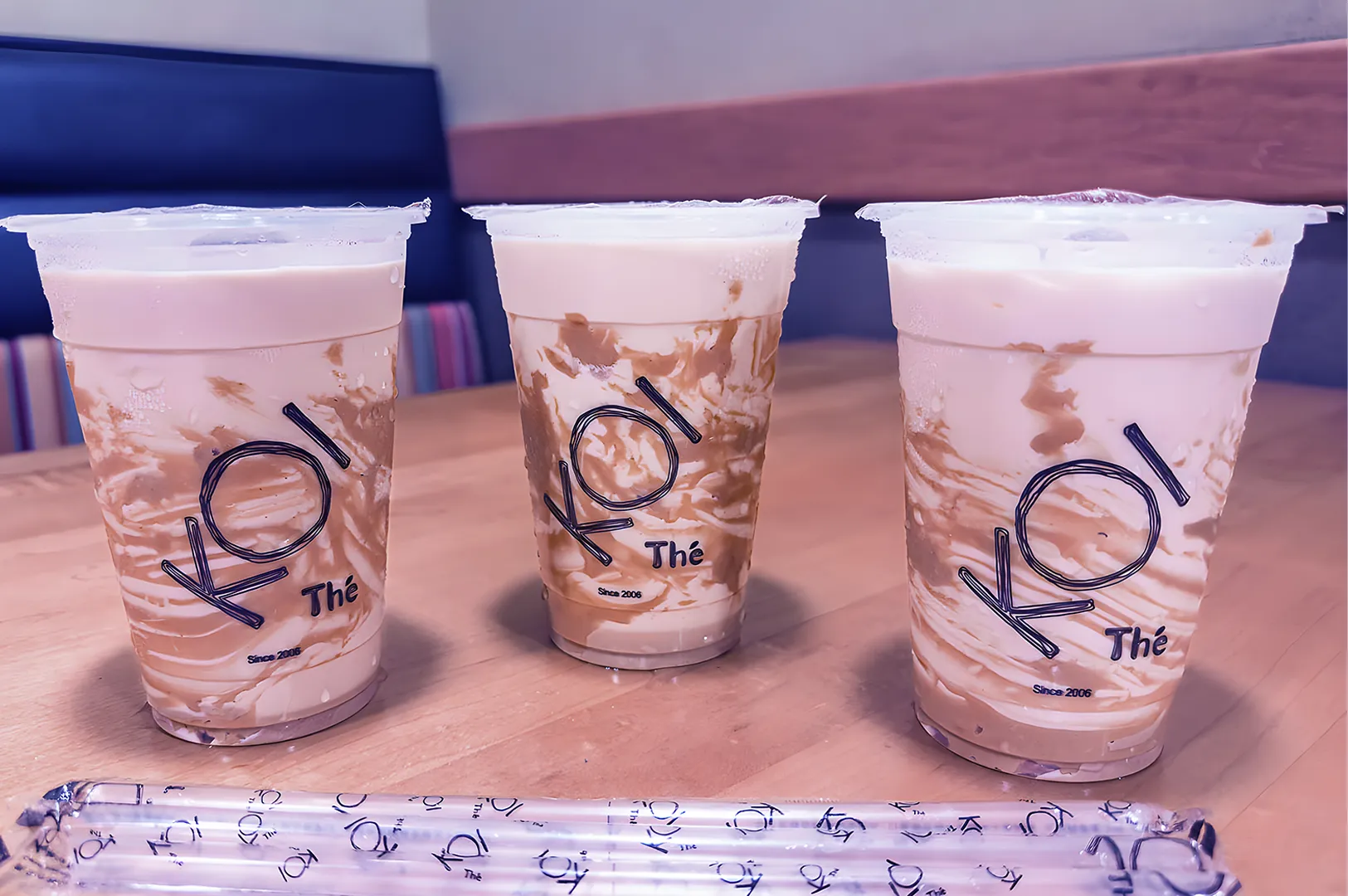 Three iced milk teas in transparent cups on a wooden table, labeled "KOI Thé." Swirled caramel is visible, creating a rich, inviting look.