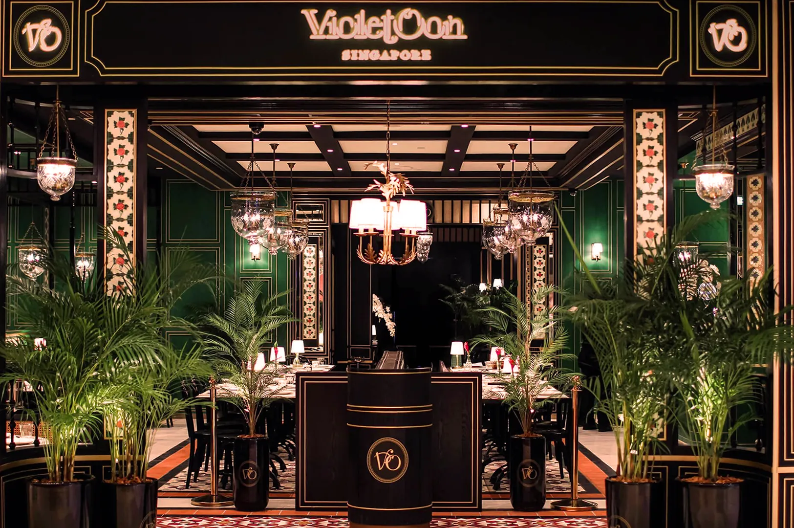 Elegant restaurant entrance with lush green plants, ornate black and gold decor, chandeliers, and a welcoming ambiance. Sign reads "Violet Oon Singapore."