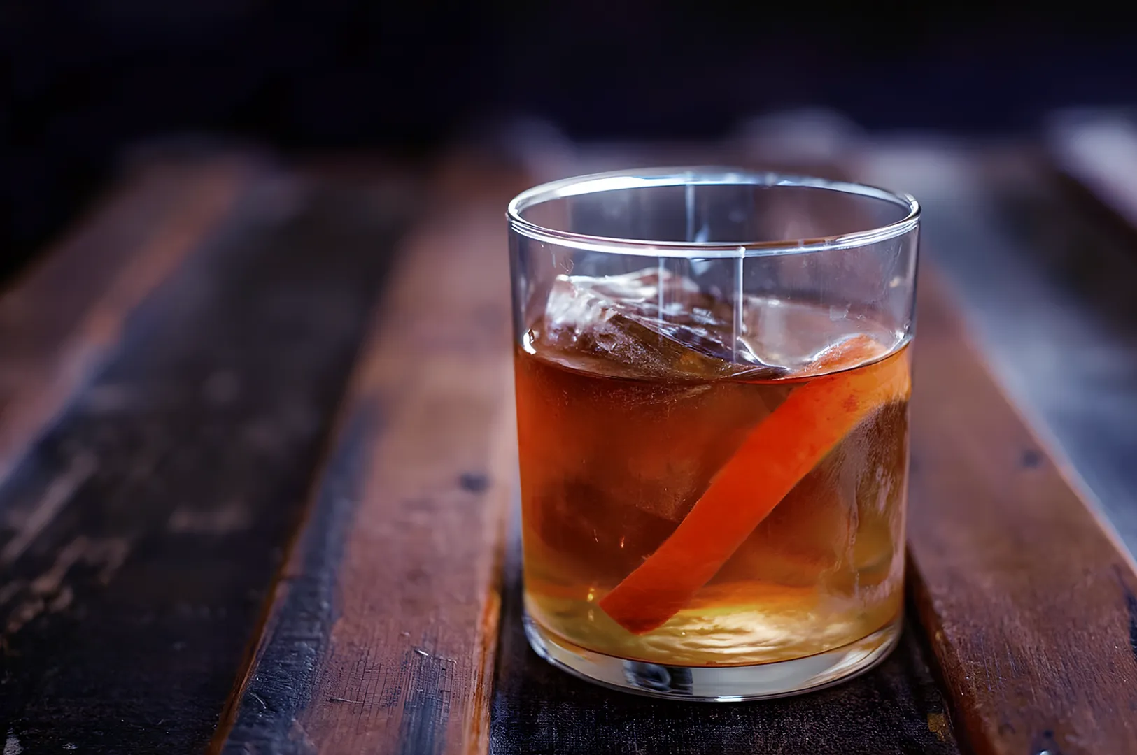 A glass of whiskey with ice and an orange peel sits on a rustic wooden table. The setting is moody and warm, evoking a cozy, relaxed atmosphere.
