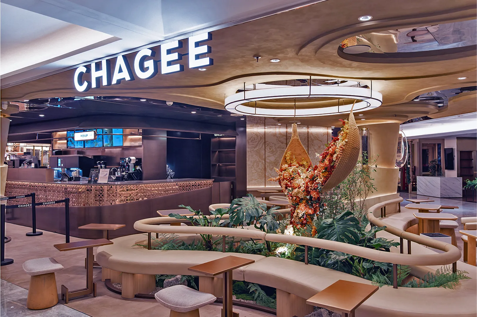 Modern tea shop interior with "CHAGEE" sign. Features curved seating, wooden tables, lush greenery, and a decorative floral arrangement in warm tones.