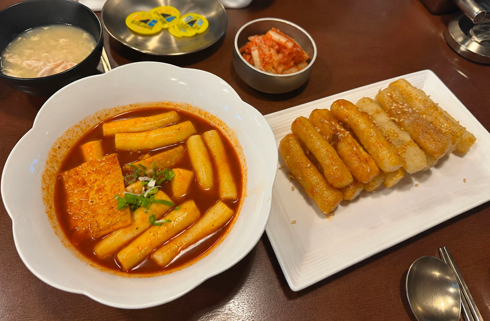 A wooden table features a bowl of spicy tteokbokki, a plate of pan-fried rice cakes, and a side of kimchii