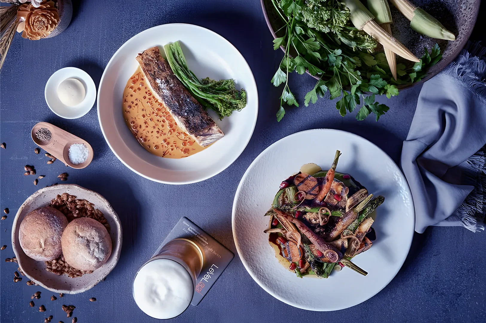 Gourmet meal on a dark table: grilled fish with sauce, roasted vegetables, bread rolls, a foamy drink, and fresh herbs. Elegant and sophisticated setting.