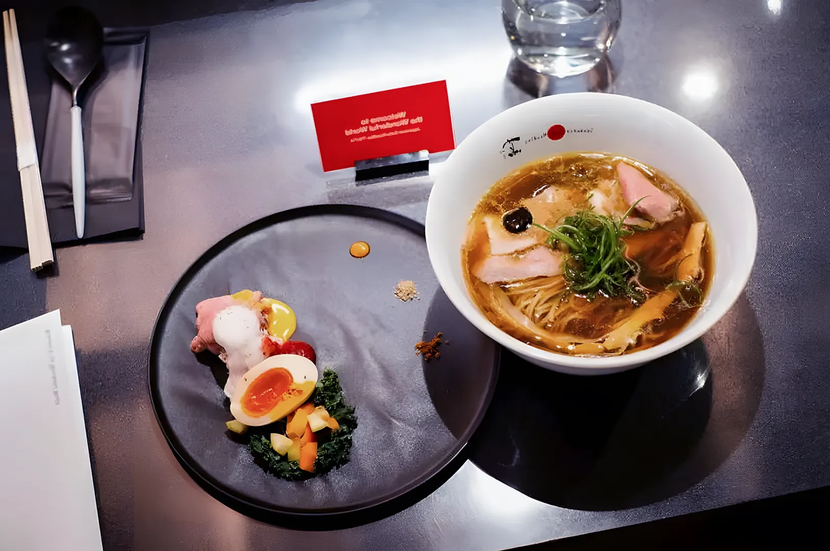 A bowl of ramen with sliced pork, green onions, and seaweed beside a plate of assorted garnishes, including a soft-boiled egg and vegetables, on a sleek, reflective table.