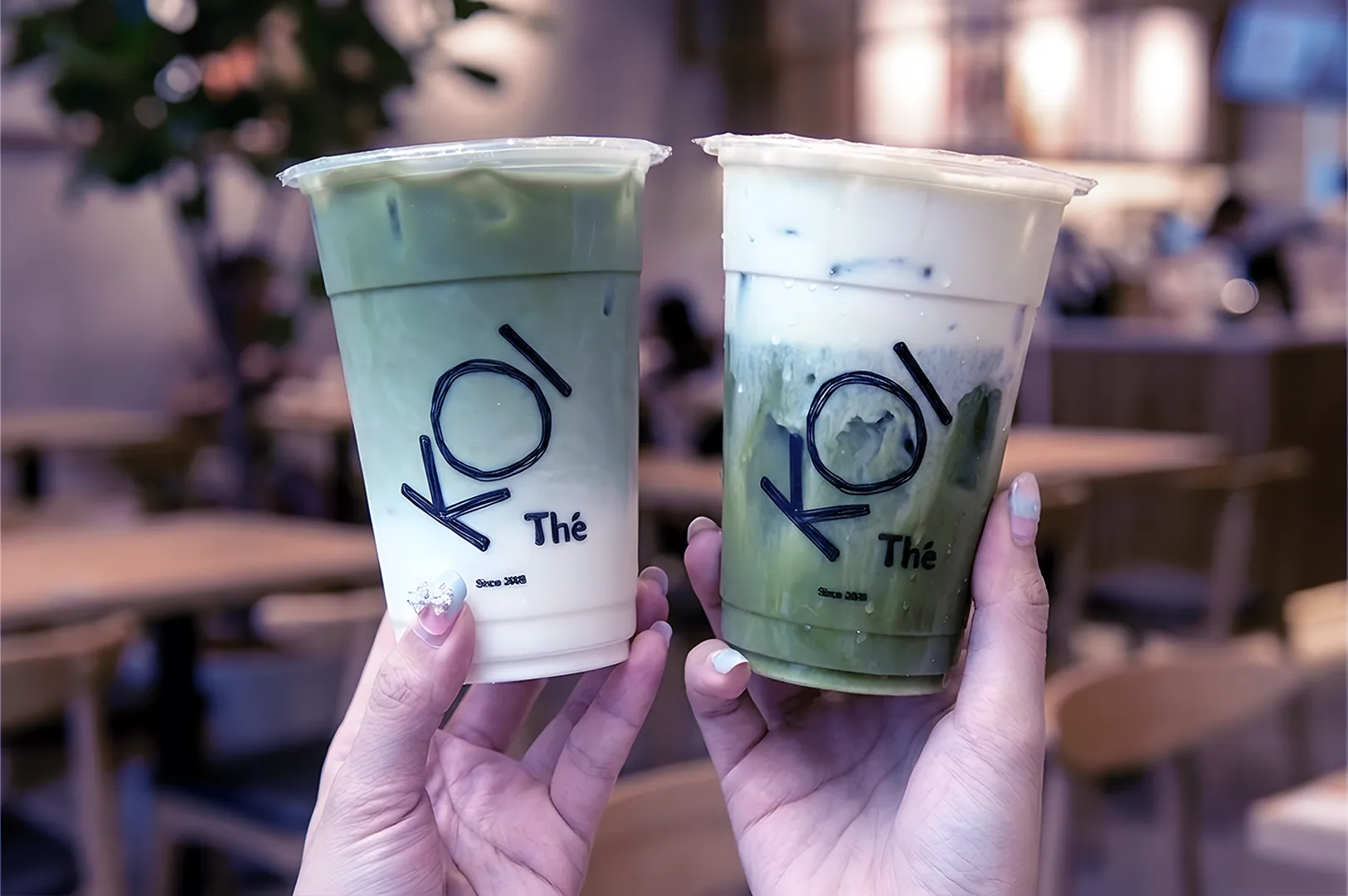 Two hands holding iced green tea drinks in clear cups from KOI Thé, inside a café with blurred wooden tables and chairs in the background.