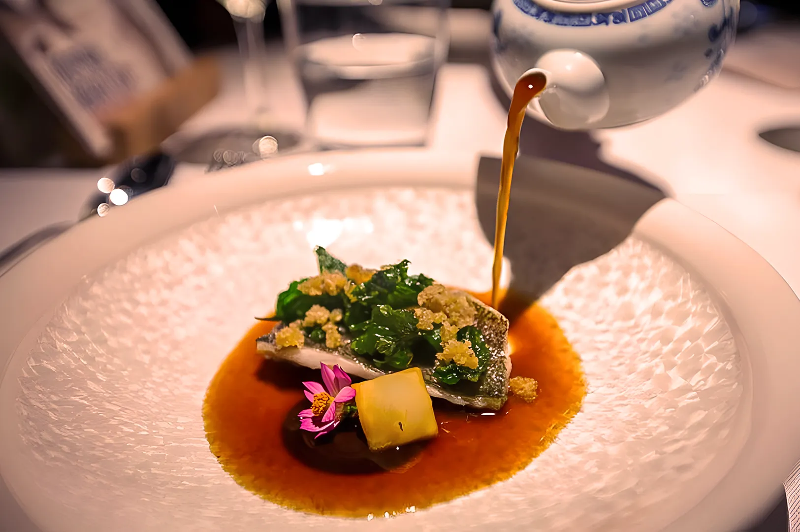 Elegant dish with fish, greens, and crumbled toppings served in a patterned bowl; sauce poured from a teapot; garnished with a pink flower, creating a gourmet presentation.