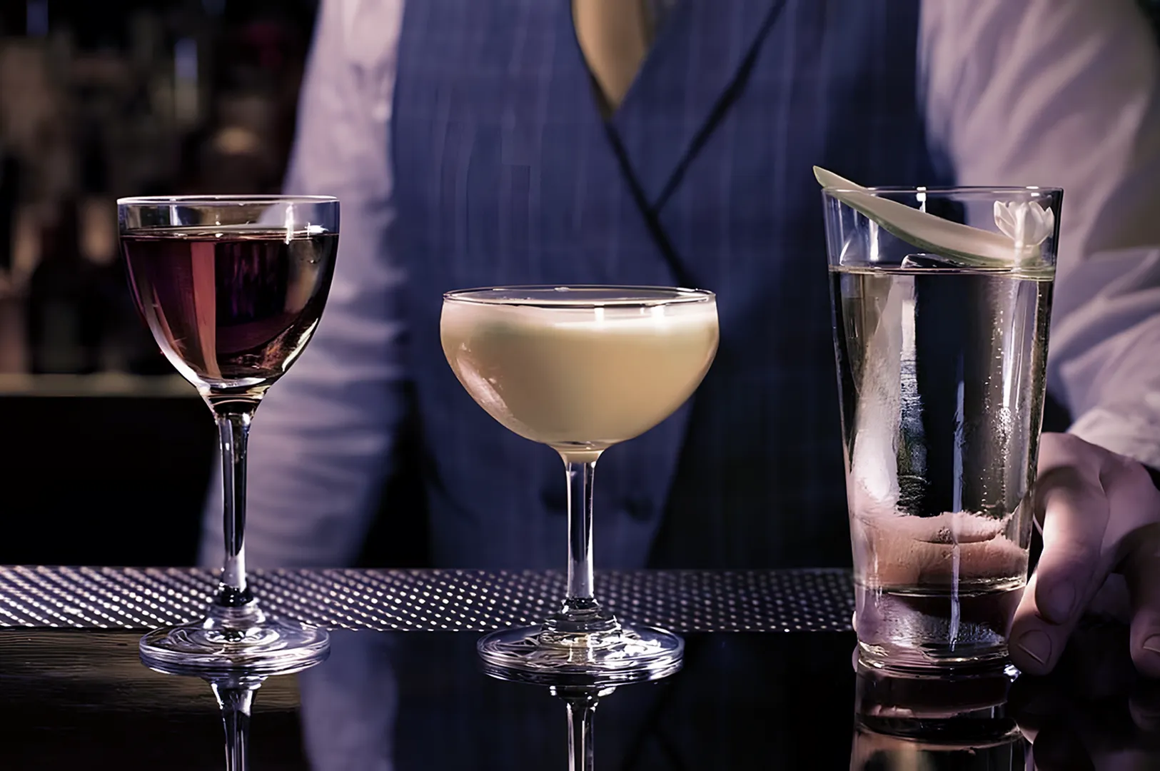 Three cocktails on a bar: a deep red drink, a creamy cocktail, and a clear drink with garnish. A person in a blue vest stands behind, creating an elegant atmosphere.
