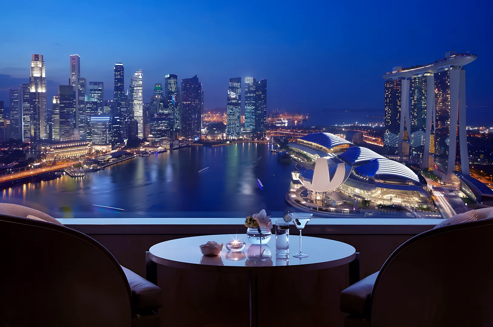 Elegant rooftop view of a cityscape at dusk, featuring illuminated skyscrapers and a waterfront. Two chairs and a table with drinks in the foreground.