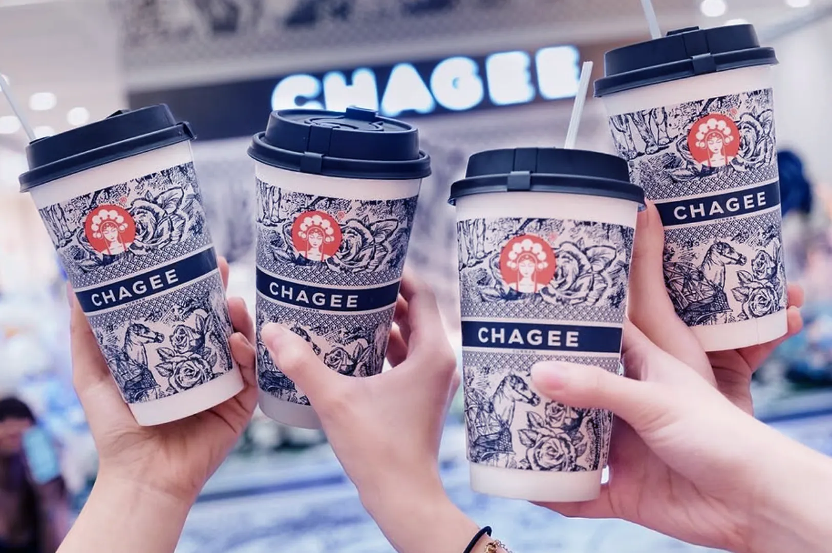 Four hands hold up Chagee cups with intricate floral patterns and navy lids, set against a blurred background of a bright store sign, conveying a sense of celebration.