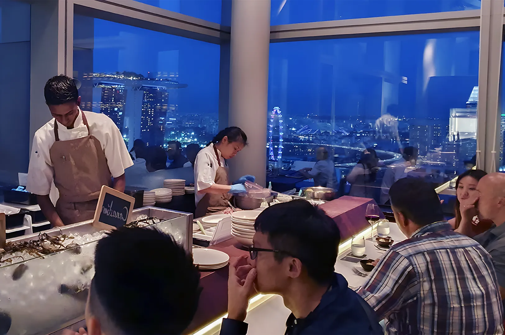 A lively rooftop restaurant with chefs preparing dishes behind a counter, diners engaging at tables. The large windows reveal a stunning city skyline at night.