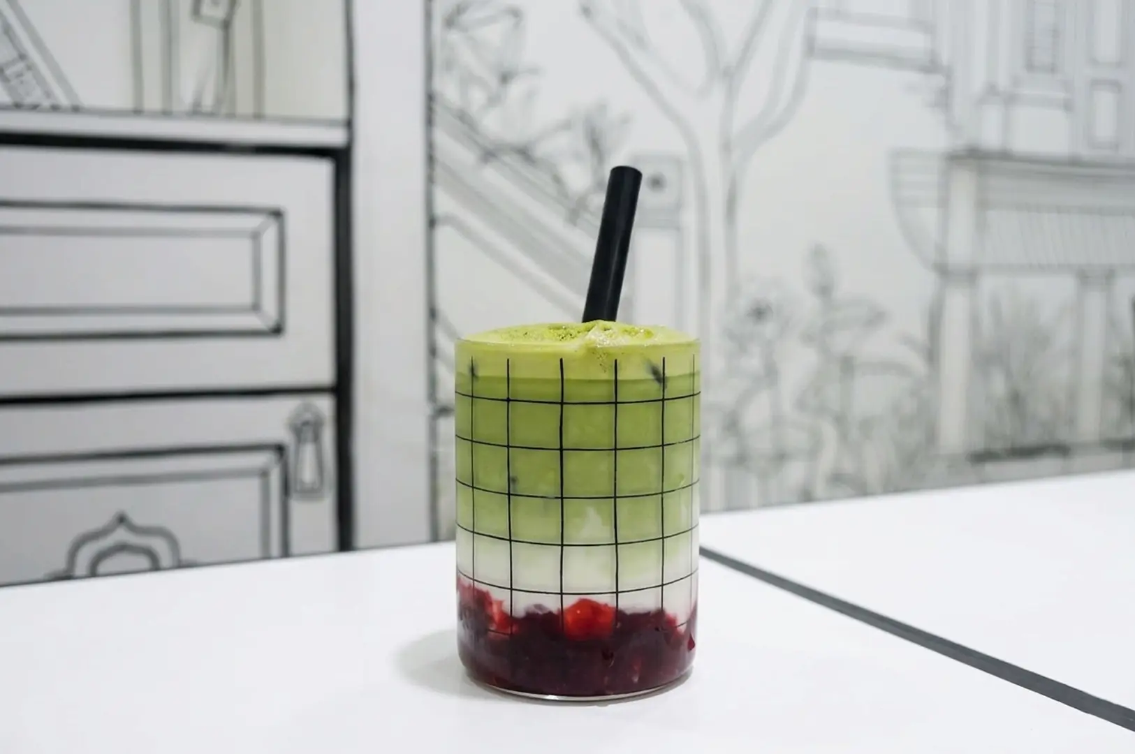 Layered matcha drink with red bean base in checkered glass with black straw against 2D line-drawn cafe background