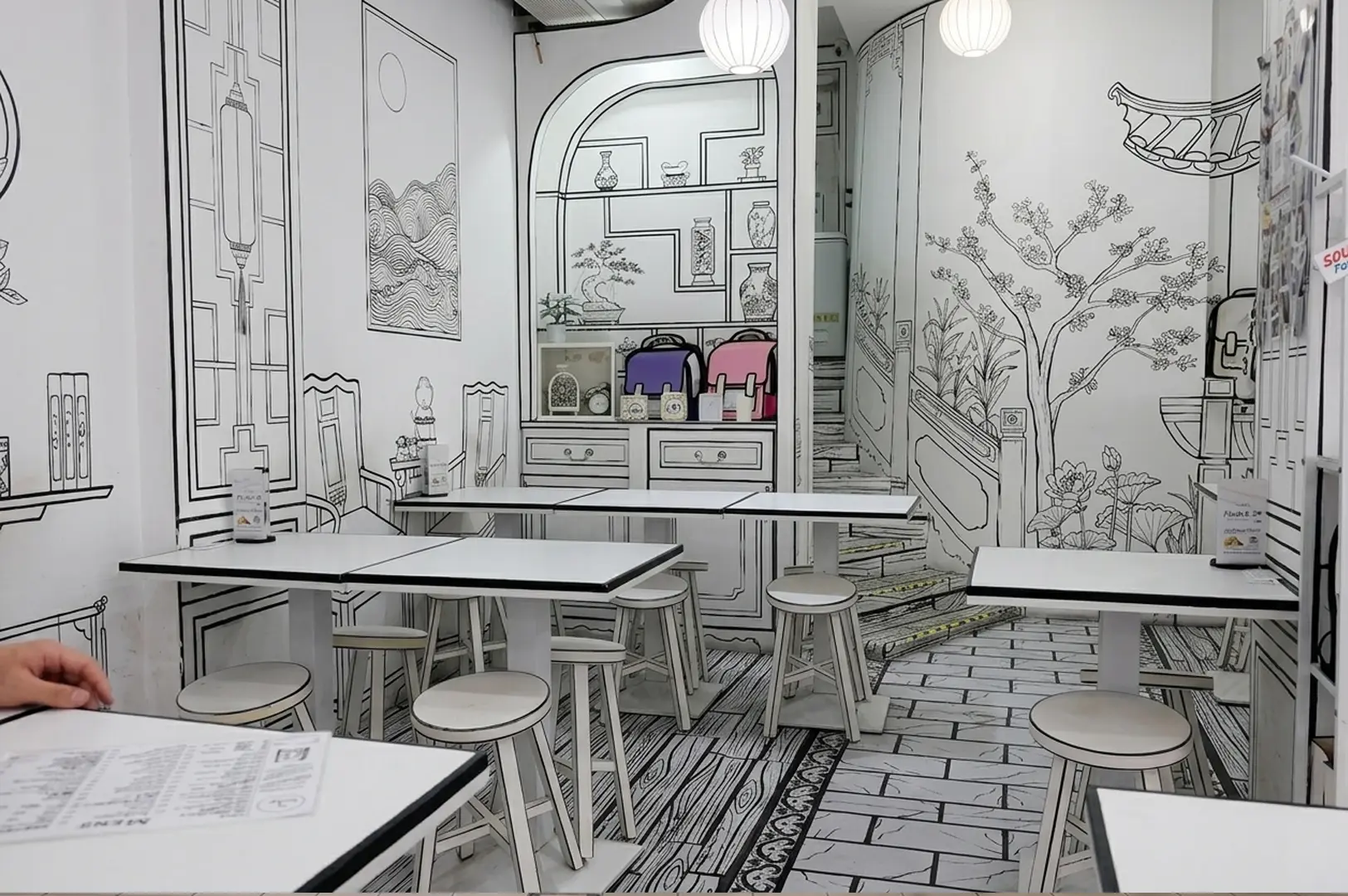 Black and white 2D line-drawn cafe interior with tables, stools, and comic book-style wall murals creating optical illusion