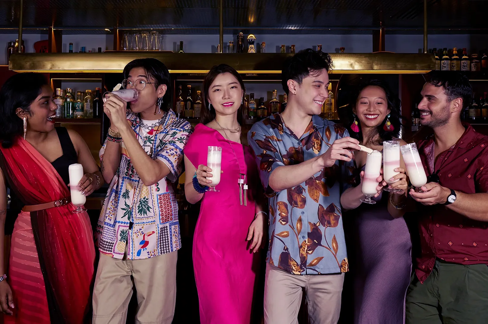 A group of six people at a bar, smiling and holding tall glasses with white beverages. The setting is vibrant, conveying joy and celebration.