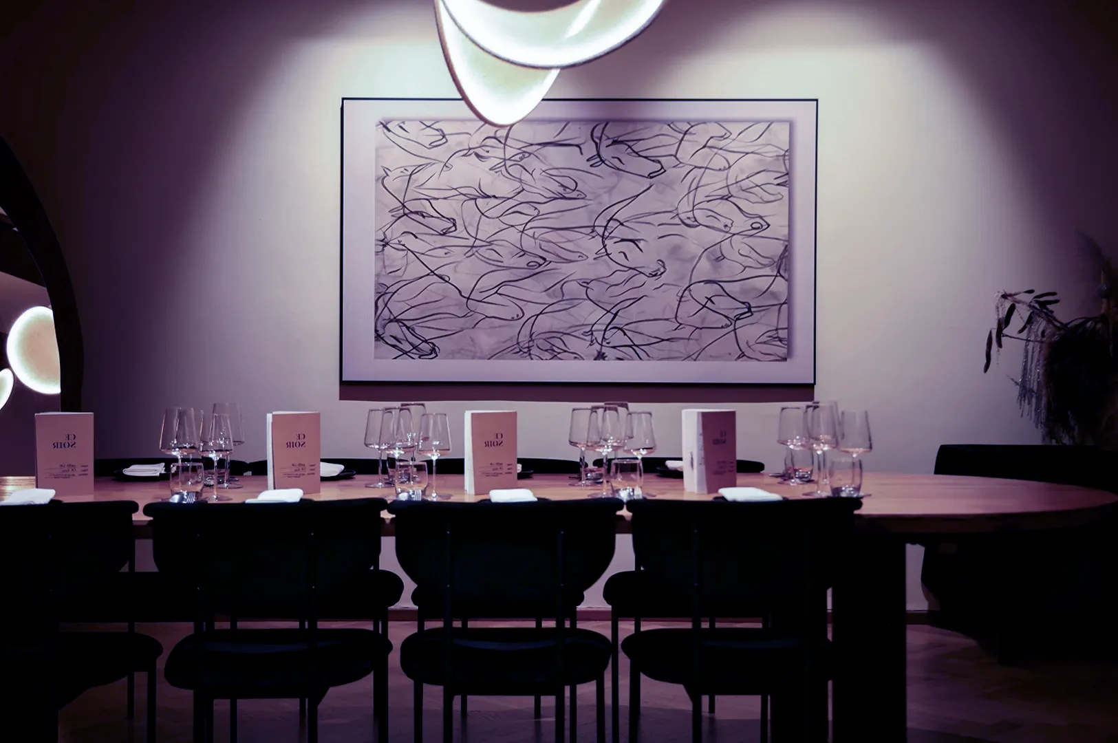 A stylish dining room with a long wooden table set for a meal. Elegant wine glasses, neatly placed menus, and modern art on the wall create a sophisticated ambiance.