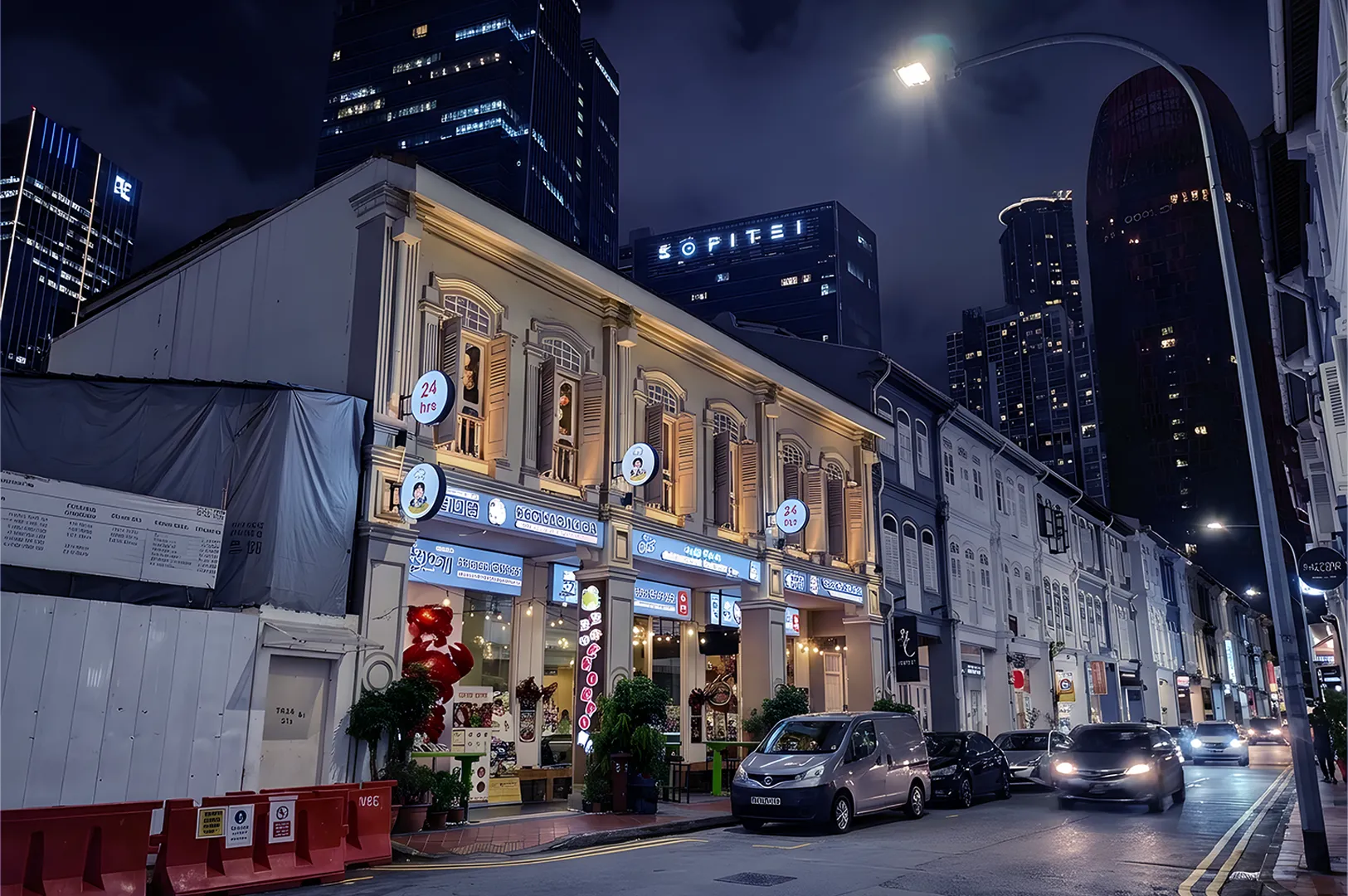 Night street scene with a row of illuminated shophouses and modern skyscrapers in the background. Cars drive by, adding a lively urban feel.